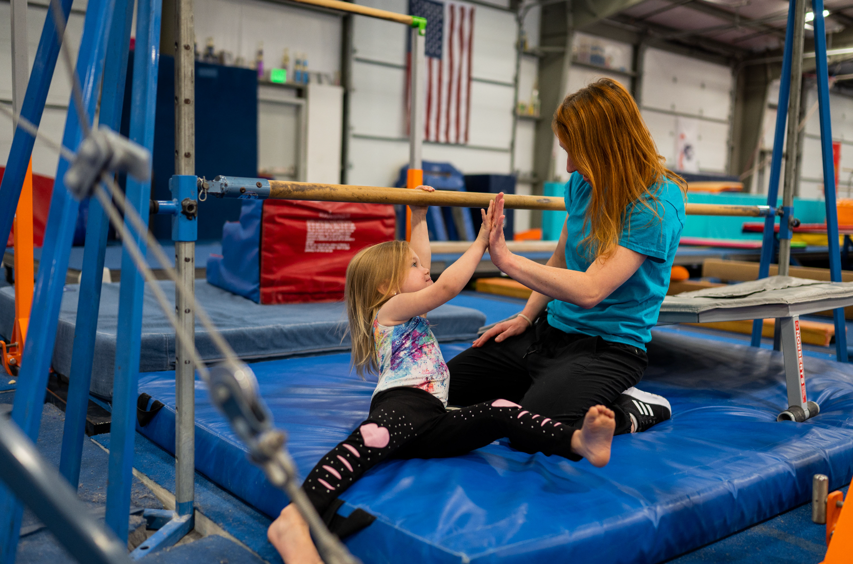 A young girl and woman high-five in a gymnastics gym. The girl is on a mat near bars.