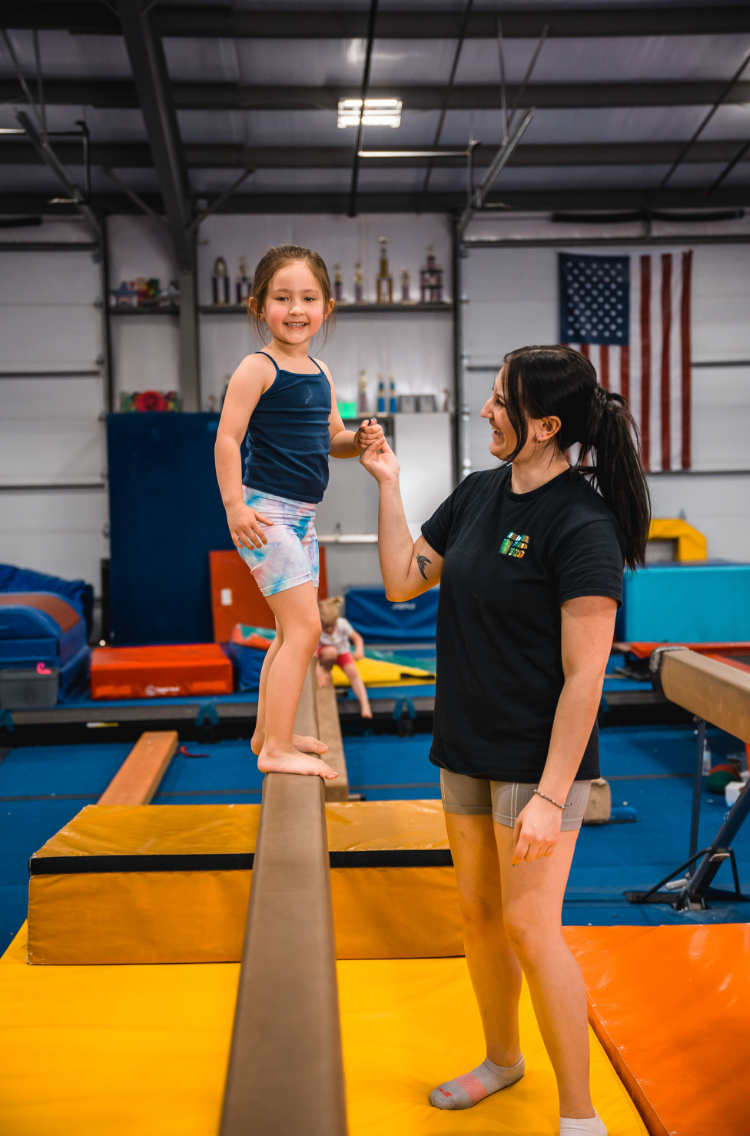 A young girl balances on a beam with a coach assisting her in a gym. Both smile with excitement; an American flag hangs in the background.