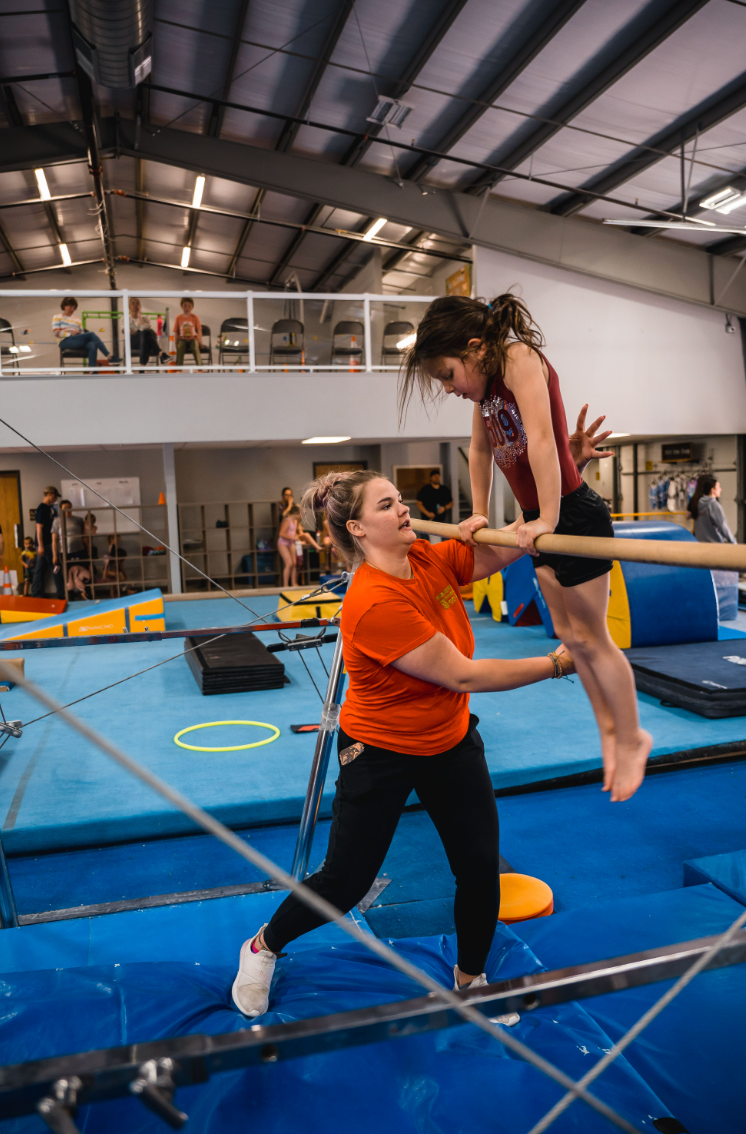 A young gymnast being assisted by a coach on uneven bars in a gymnasium. The coach is wearing an orange shirt and the gymnast is wearing a leotard.