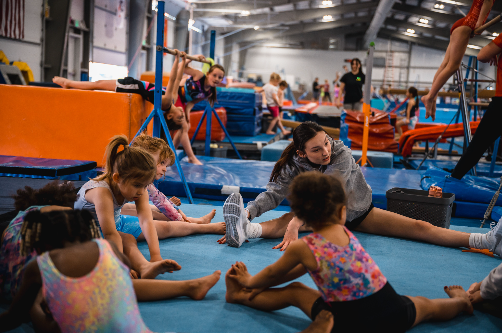 Young gymnasts stretching on a blue mat in a gym, with a coach assisting. Other gymnasts are practicing on equipment.