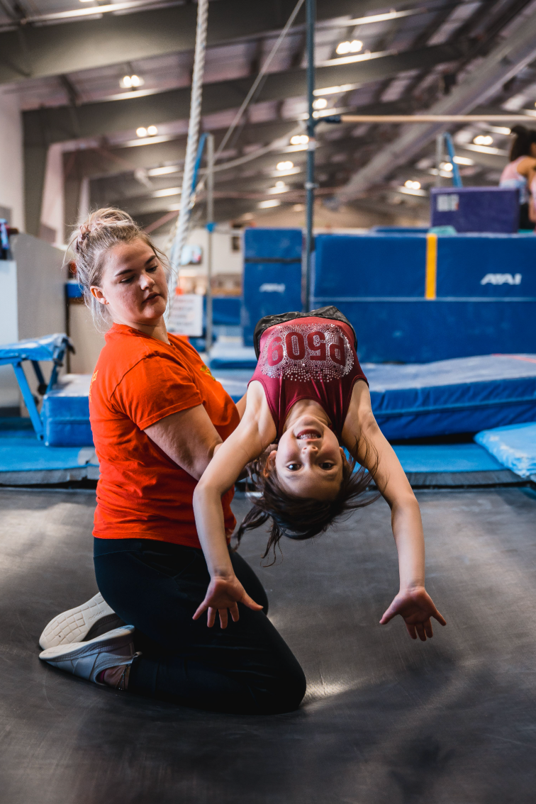A gymnastics instructor assists a young girl with a backbend on a mat in a gym. The instructor wears orange, the girl a maroon top.