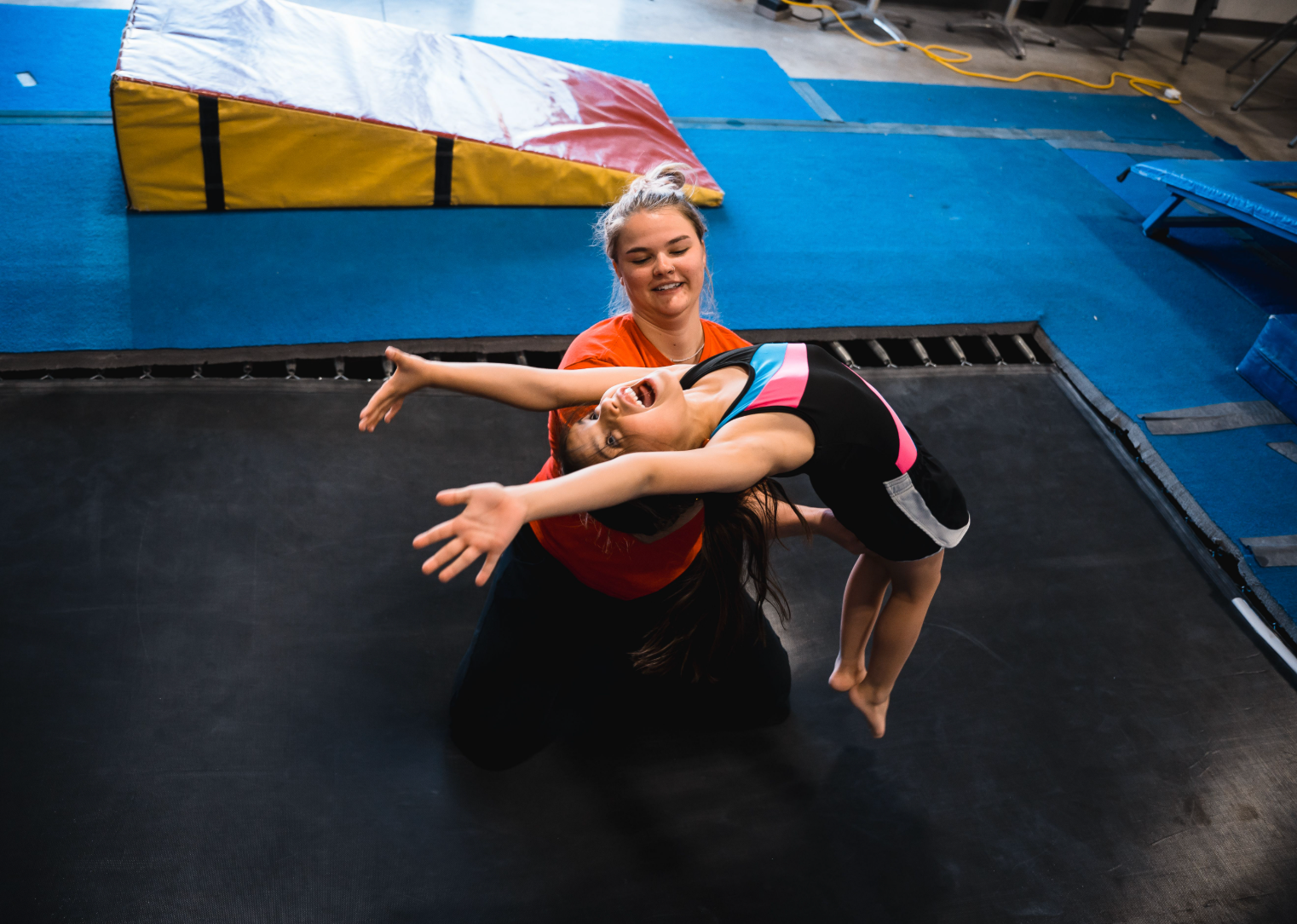 A young girl arches backward on a trampoline, assisted by a kneeling coach with arms outstretched. Blue floor, indoor setting.