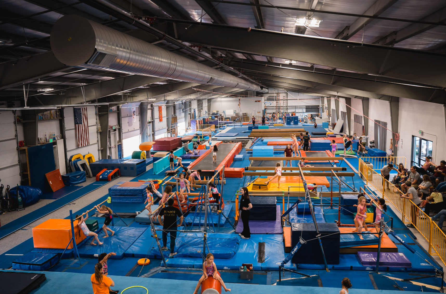 Gym with children and adults participating in a fitness class, utilizing various obstacles and mats. Blue mats and equipment fill the spacious interior.