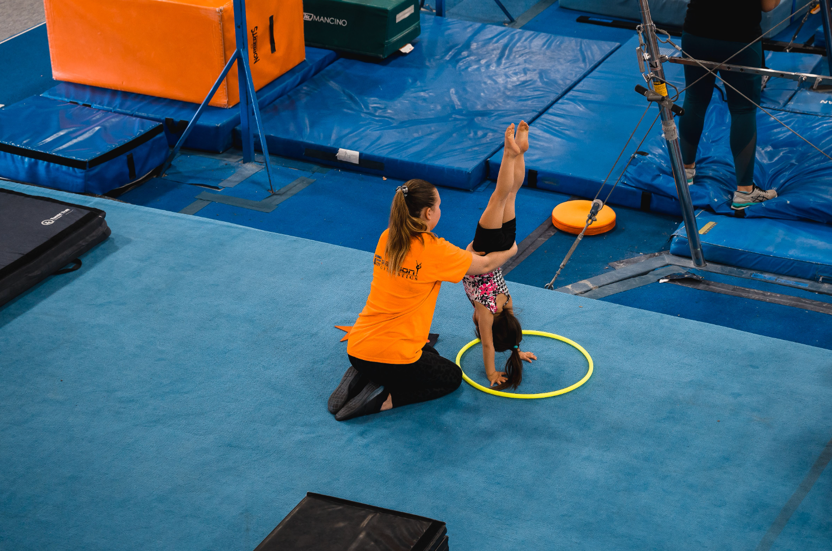 A gymnast is in a handstand position, assisted by a coach, on a blue mat. A yellow hoop is around the gymnast's hands.
