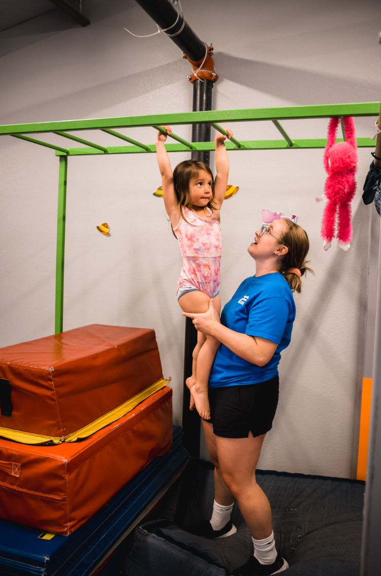 A young girl hangs from monkey bars, supported by an adult in a gym setting. The adult is smiling, while the girl is focused.