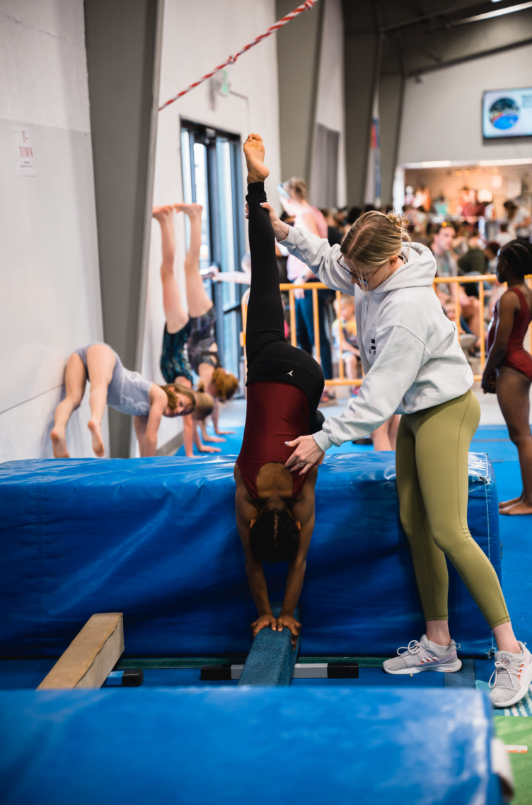 Gymnast doing a handstand on a beam, assisted by a coach. Training in a gym setting with other gymnasts practicing.