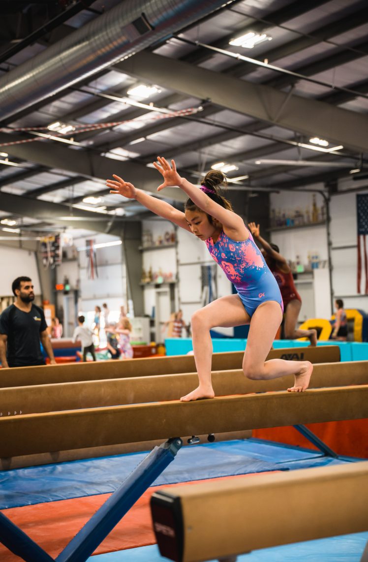 A young girl in a blue and pink leotard leaps from a balance beam in a gymnastics gym.