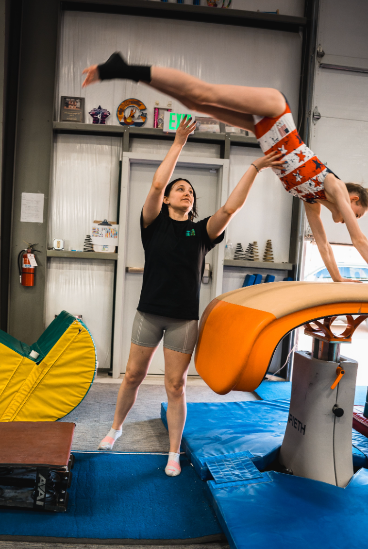 Gymnast performing vault with coach's assistance. The setting is a gym with blue mats, an orange vault, and a person in a black shirt.