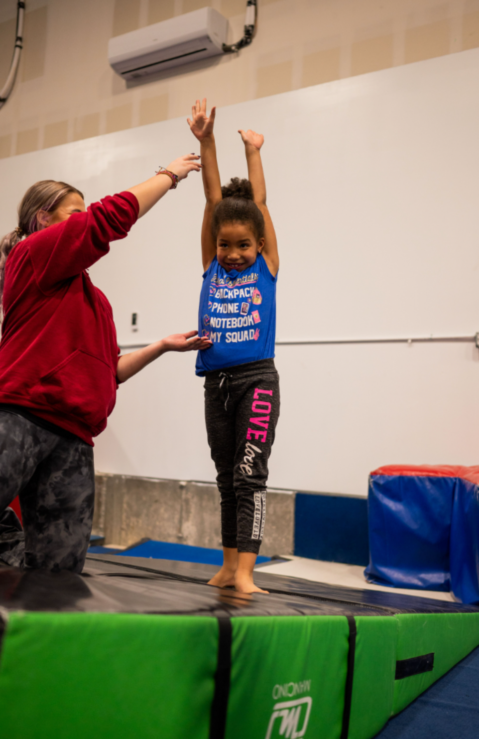 Young girl with dark skin in a gymnastics class, arms raised, with instructor guiding her. Setting is a gym.