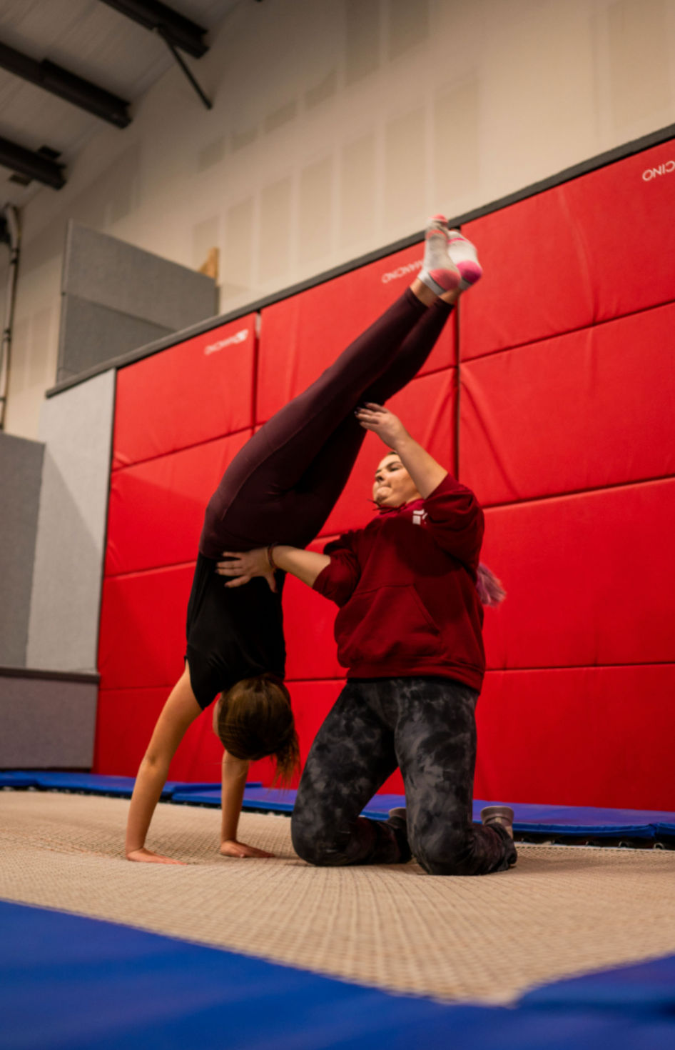Two people practicing a handstand on a trampoline. One person is upside down, being supported by the other. They are in a gym.