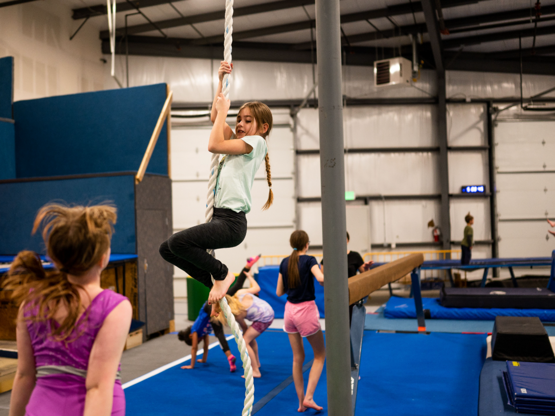A young girl climbs a rope in a gymnasium; other children are practicing gymnastics on blue mats and equipment.