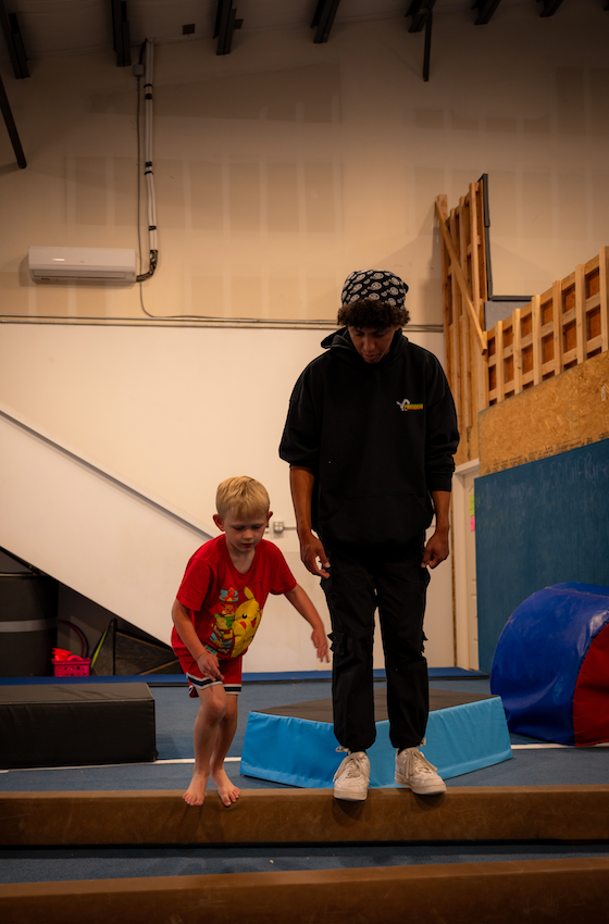 A gymnastics coach demonstrating a floor exercise to a young girl in a gym with blue mats.