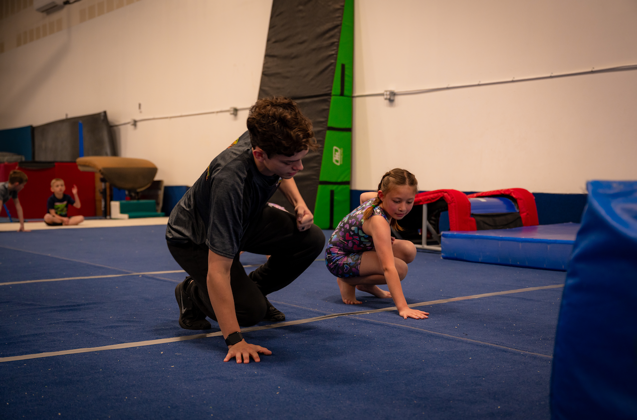 A child in a red shirt jumps down as an adult in a black hoodie stands on a beam in a gym.