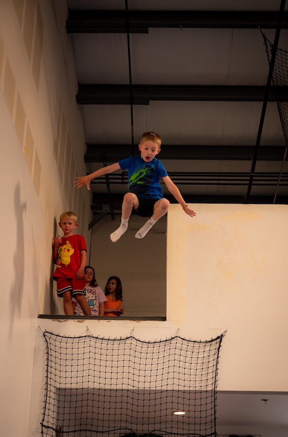 Boy jumps off platform, arms outstretched, into a netted area. Two children watch from the platform.