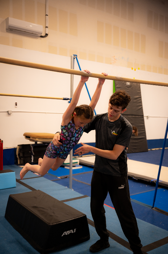Girl hanging from bar with coach's support in a gym. Black mat below, neutral background.