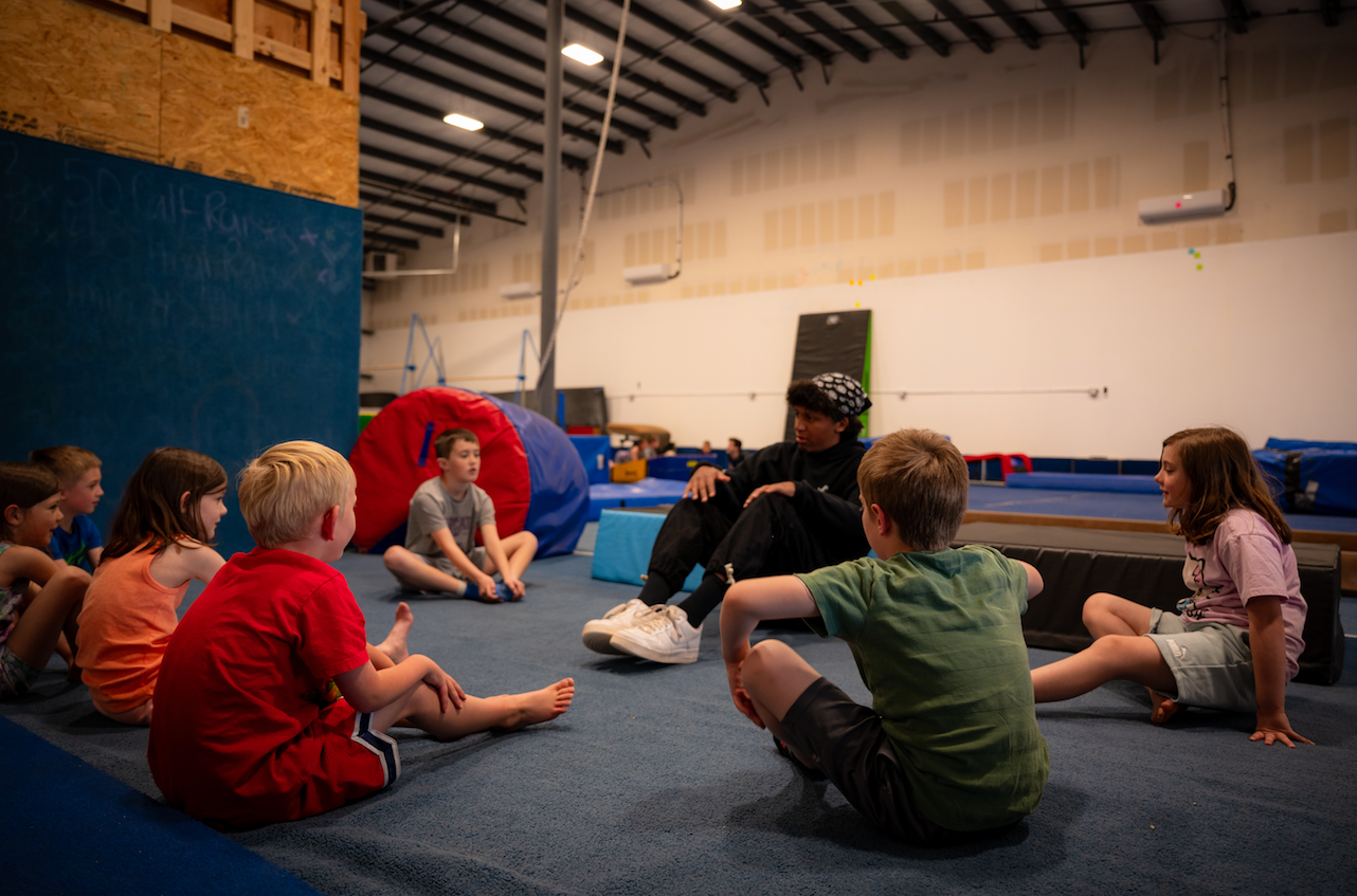 A group of children sitting on a mat listens to a person in a gym.