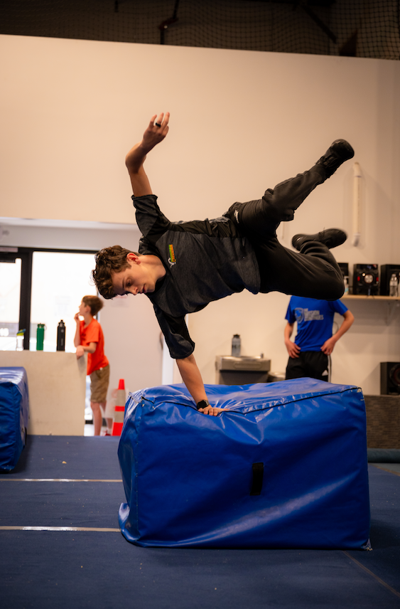 A person doing a parkour move, balancing on a blue padded block, indoor gym.