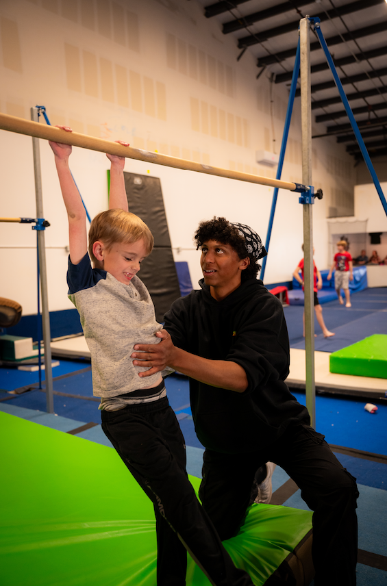 A child hangs from a bar with help from an instructor in a gymnastics studio.