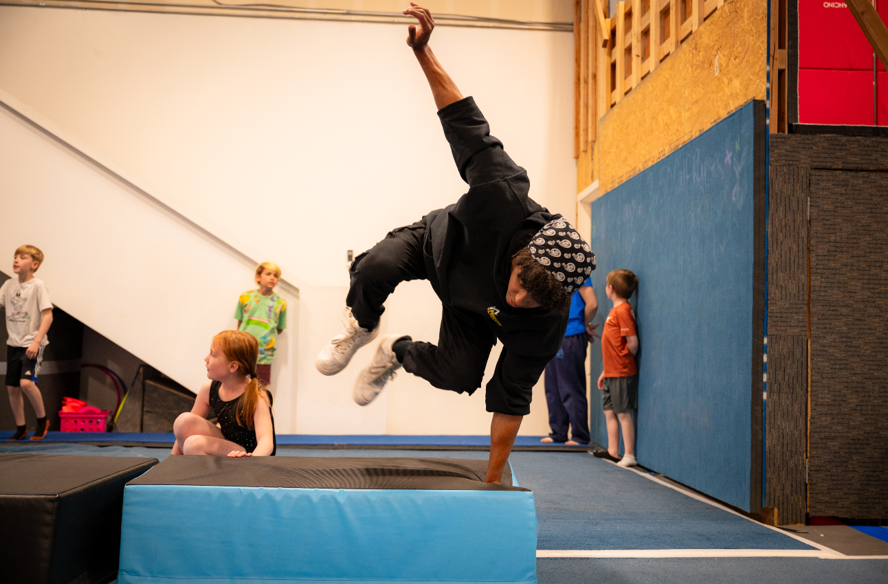 Man doing handstand on block in gym, children watch.
