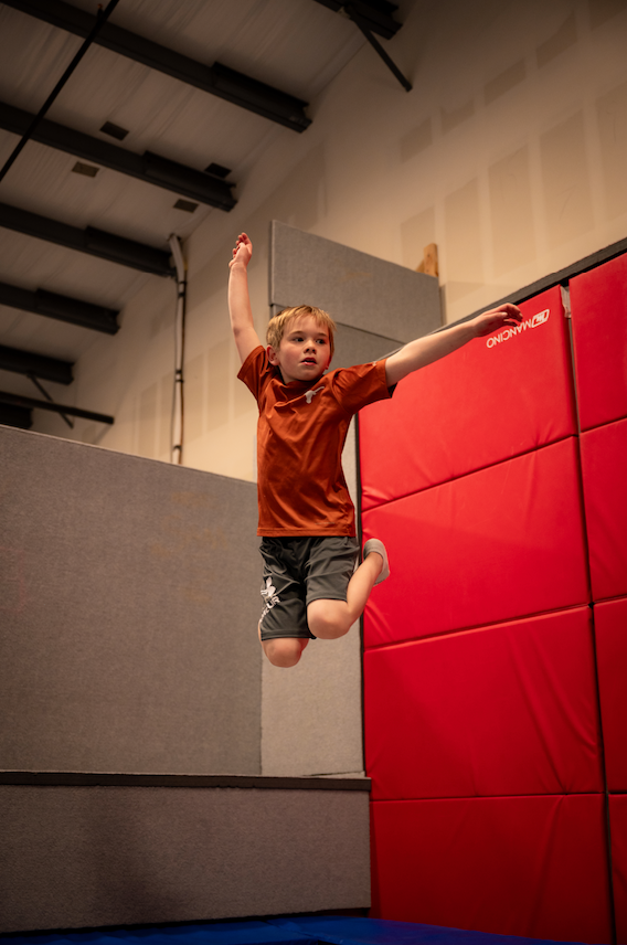 Boy leaping in the air, arms out, in a gym with red mats and a low gray wall.