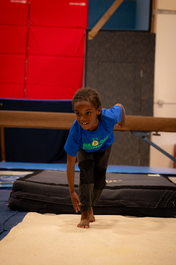 Child balancing on one foot, smiling, at a gymnastics facility. Wearing blue shirt, dark pants. In front of a balance beam.