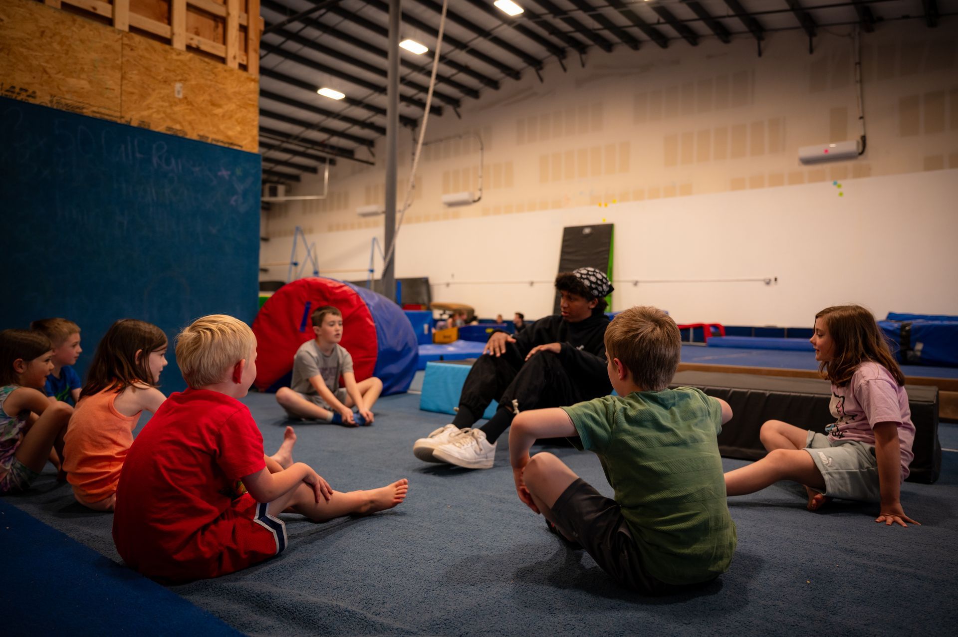 A group of children are sitting in a circle on the floor in a gym.