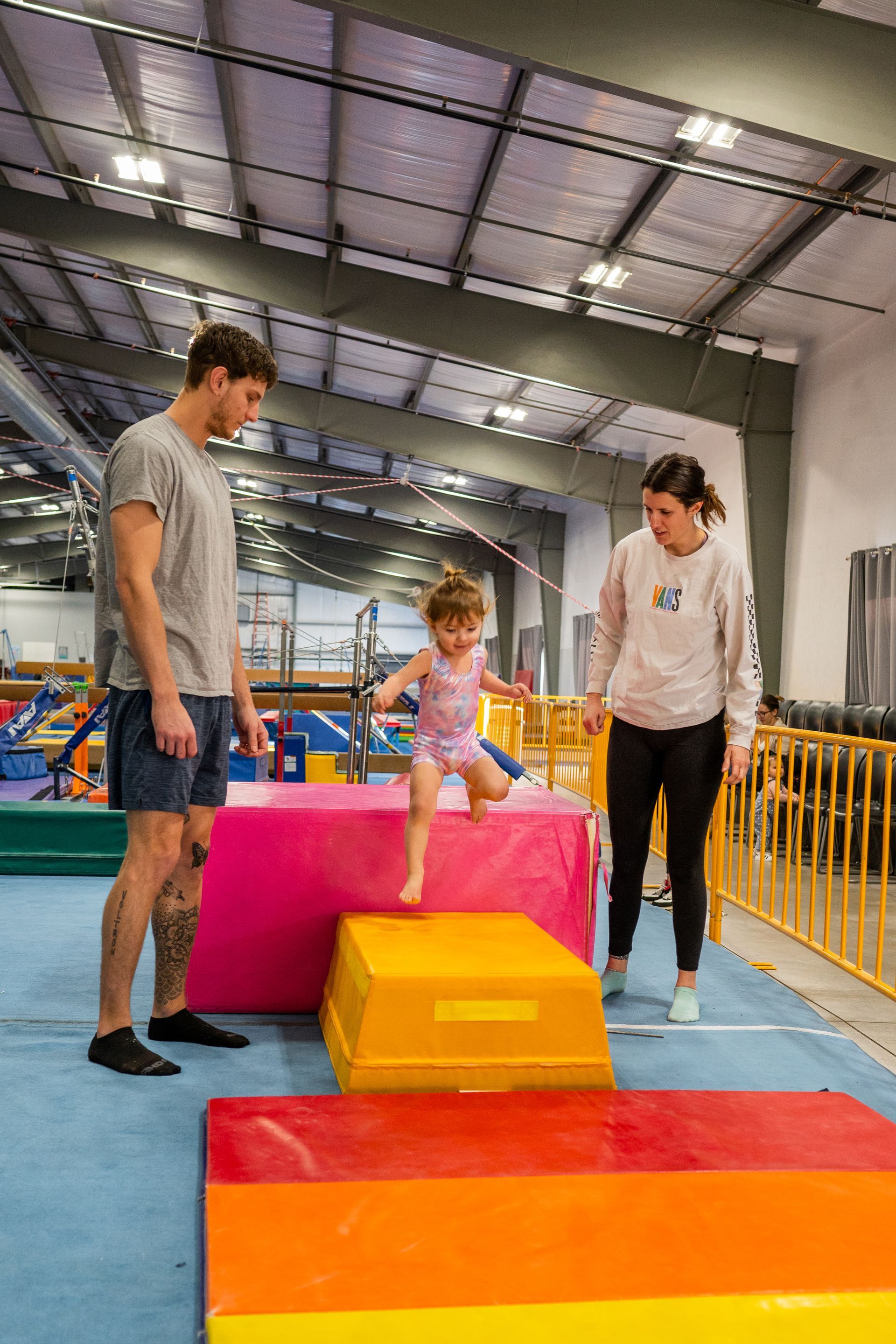 A man and woman are standing next to a little girl who is jumping over blocks in a gym.