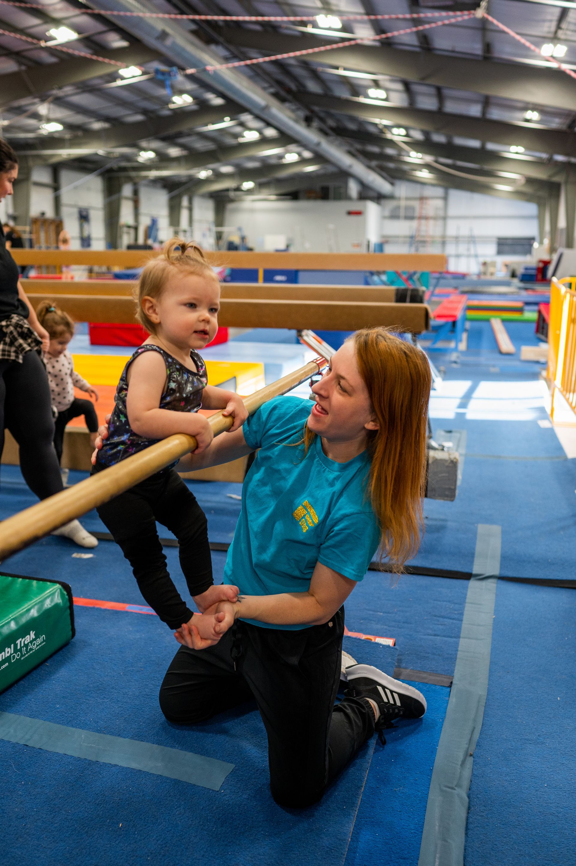 A woman is kneeling down next to a baby on a balance beam in a gym.