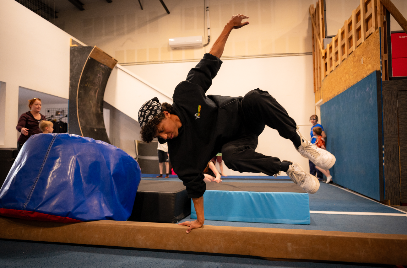 Person balances on one hand, legs in air, on a beam in a gym.