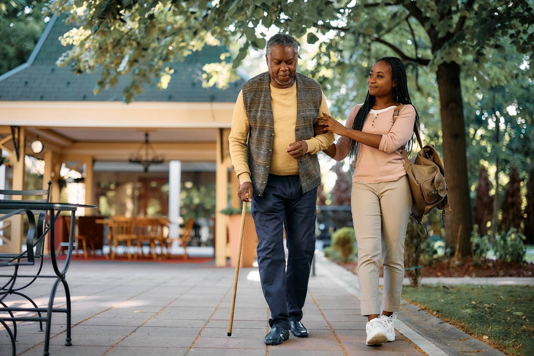 A woman is helping an elderly man with a cane walk down a sidewalk.