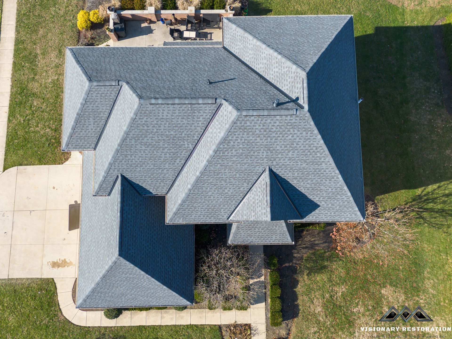 Aerial view of a gray roof on a house with multiple gables, set in a yard with concrete and grass.