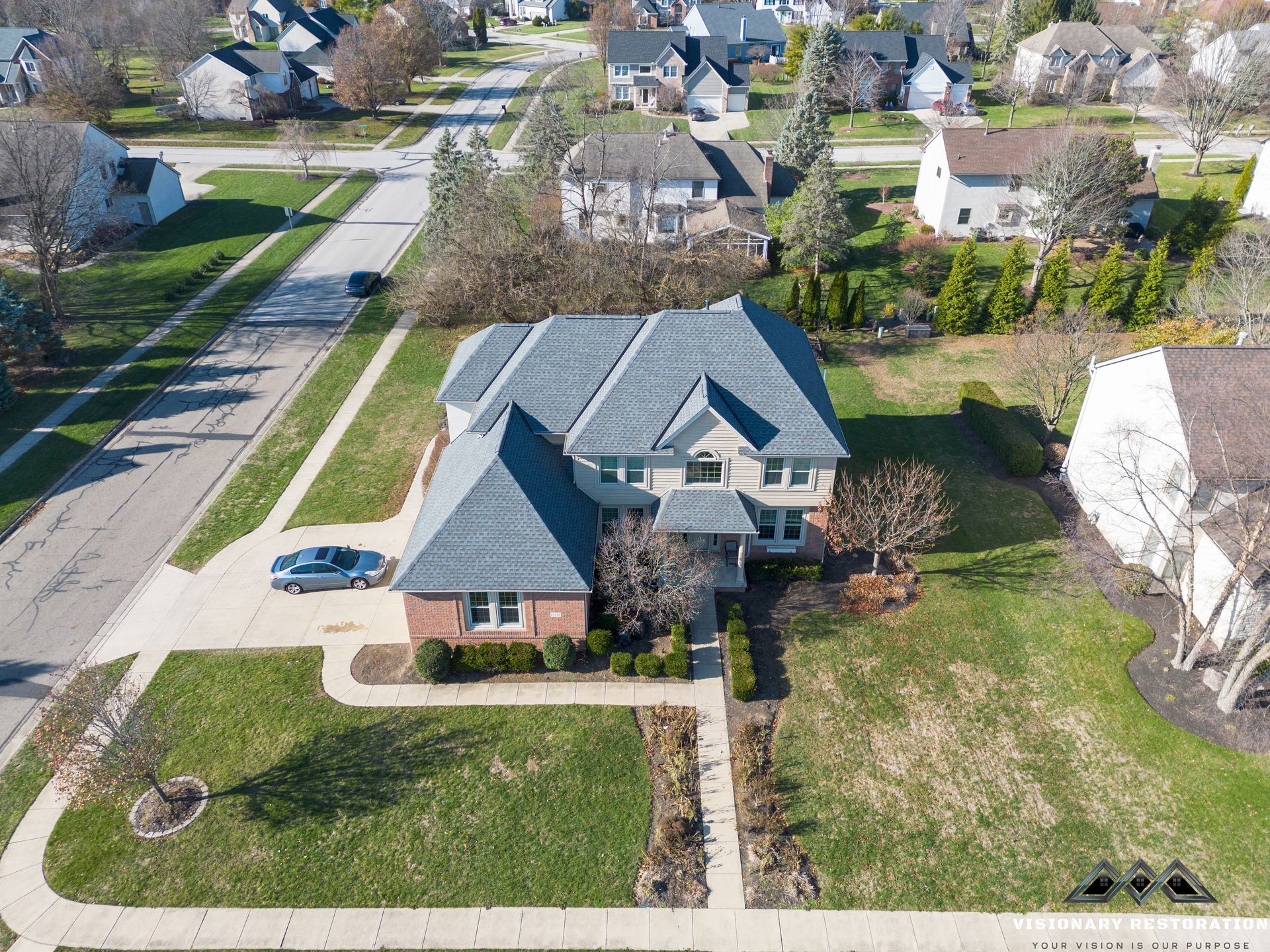 Aerial view of a two-story house with gray roof, brick and light siding, on a green lawn with a driveway and surrounding houses.