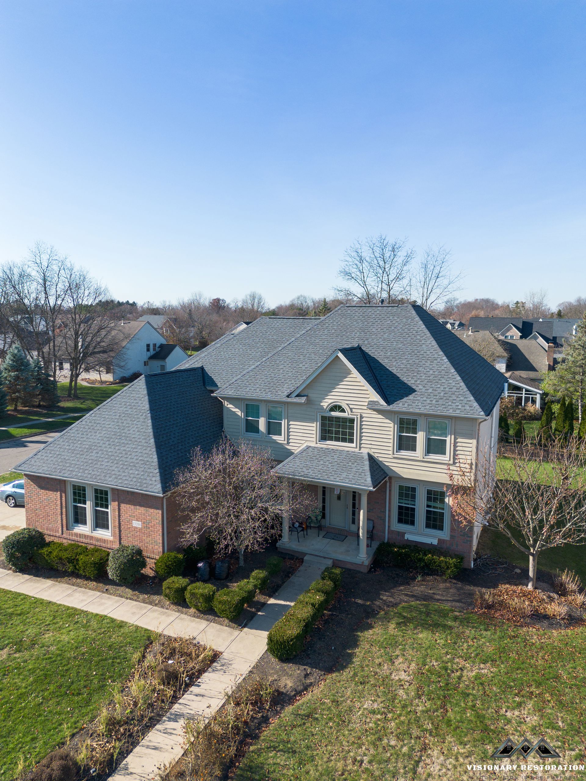 Two-story house with gray roof, red brick, and beige siding. Sunny day with trees and lawn.