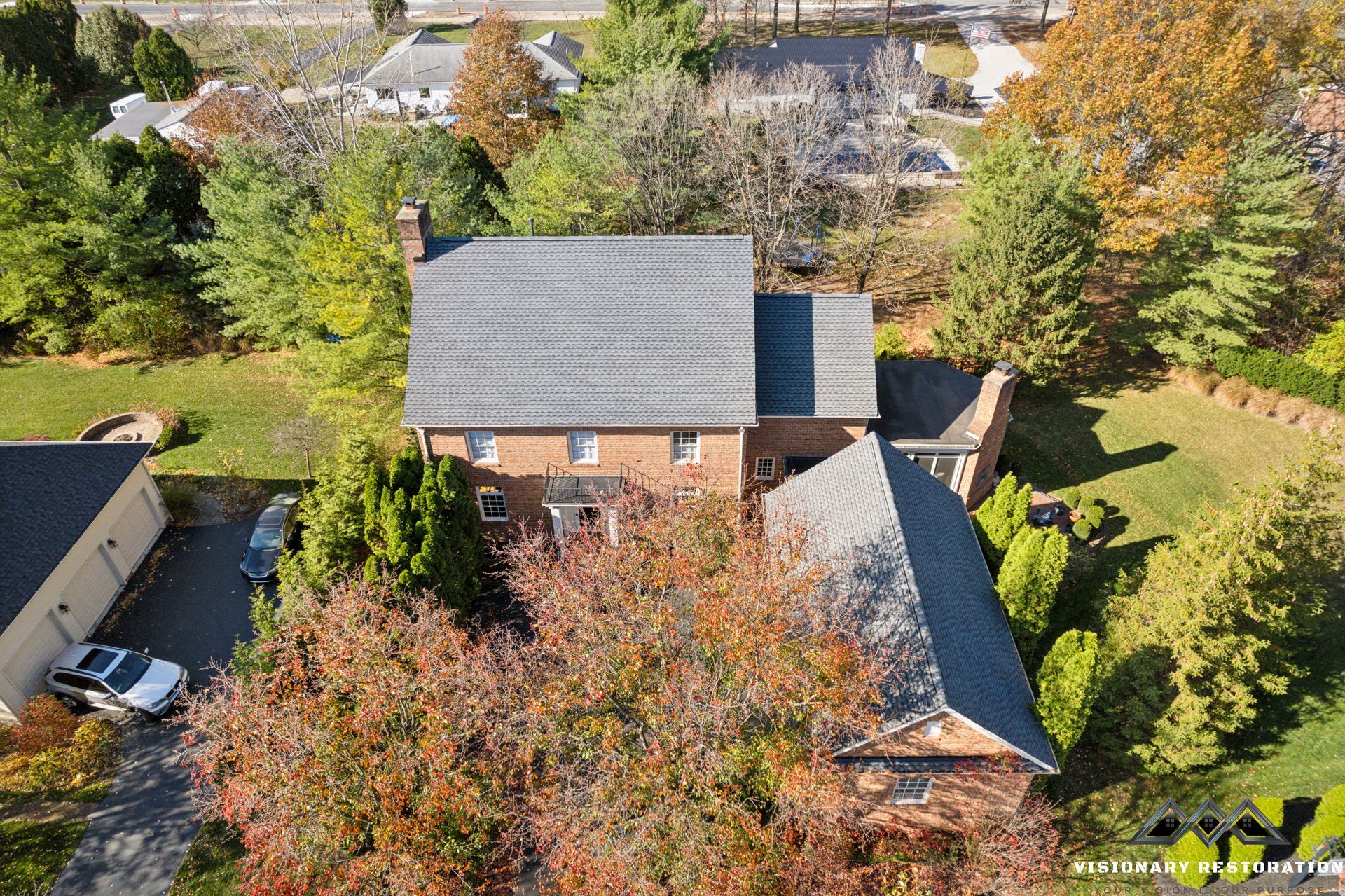 Aerial view of a two-story brick house with a gray roof, surrounded by trees with colorful fall foliage.