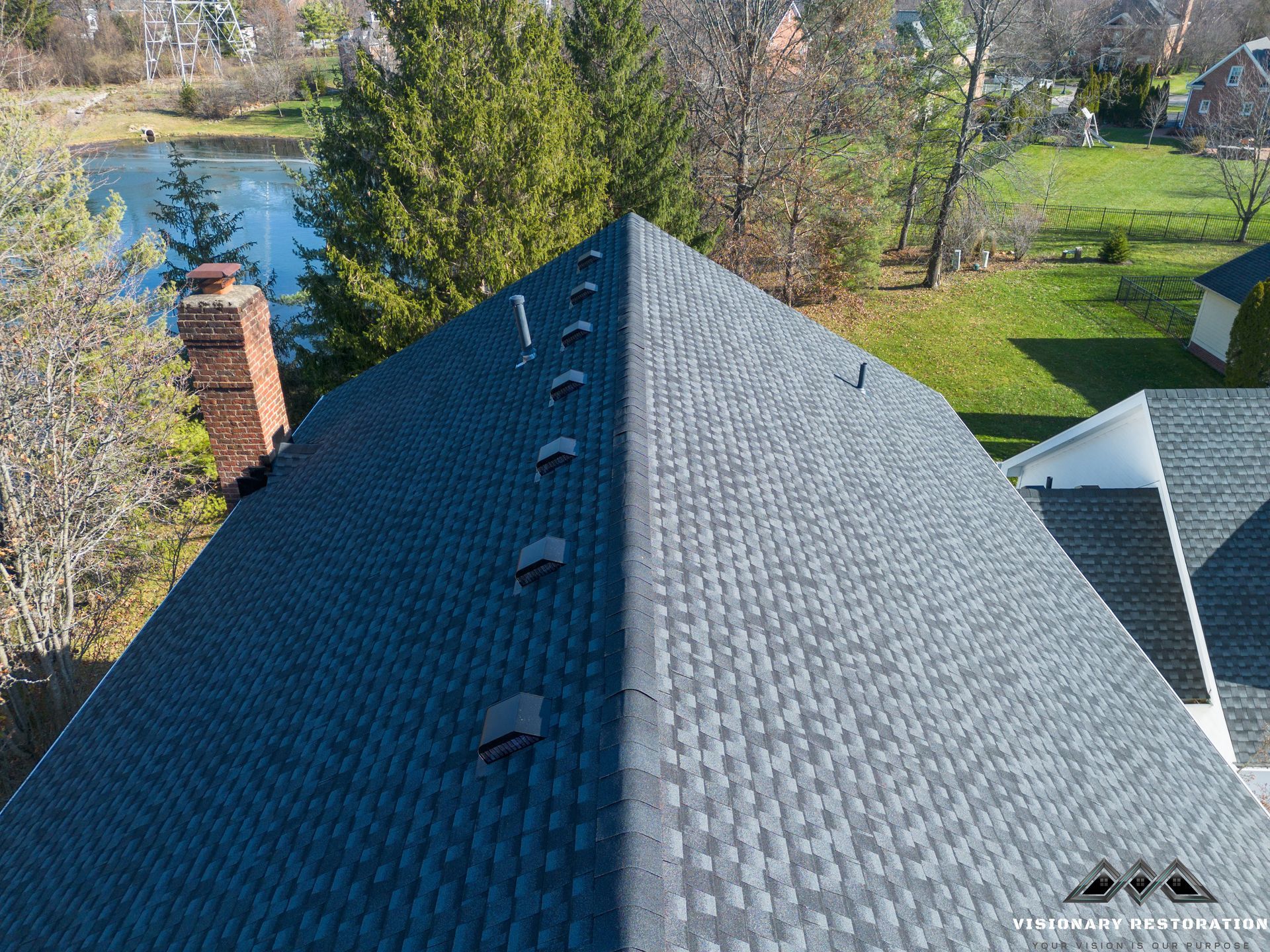 Close-up of a dark grey shingled roof with small vents along the top, overlooking a lake and green lawn.