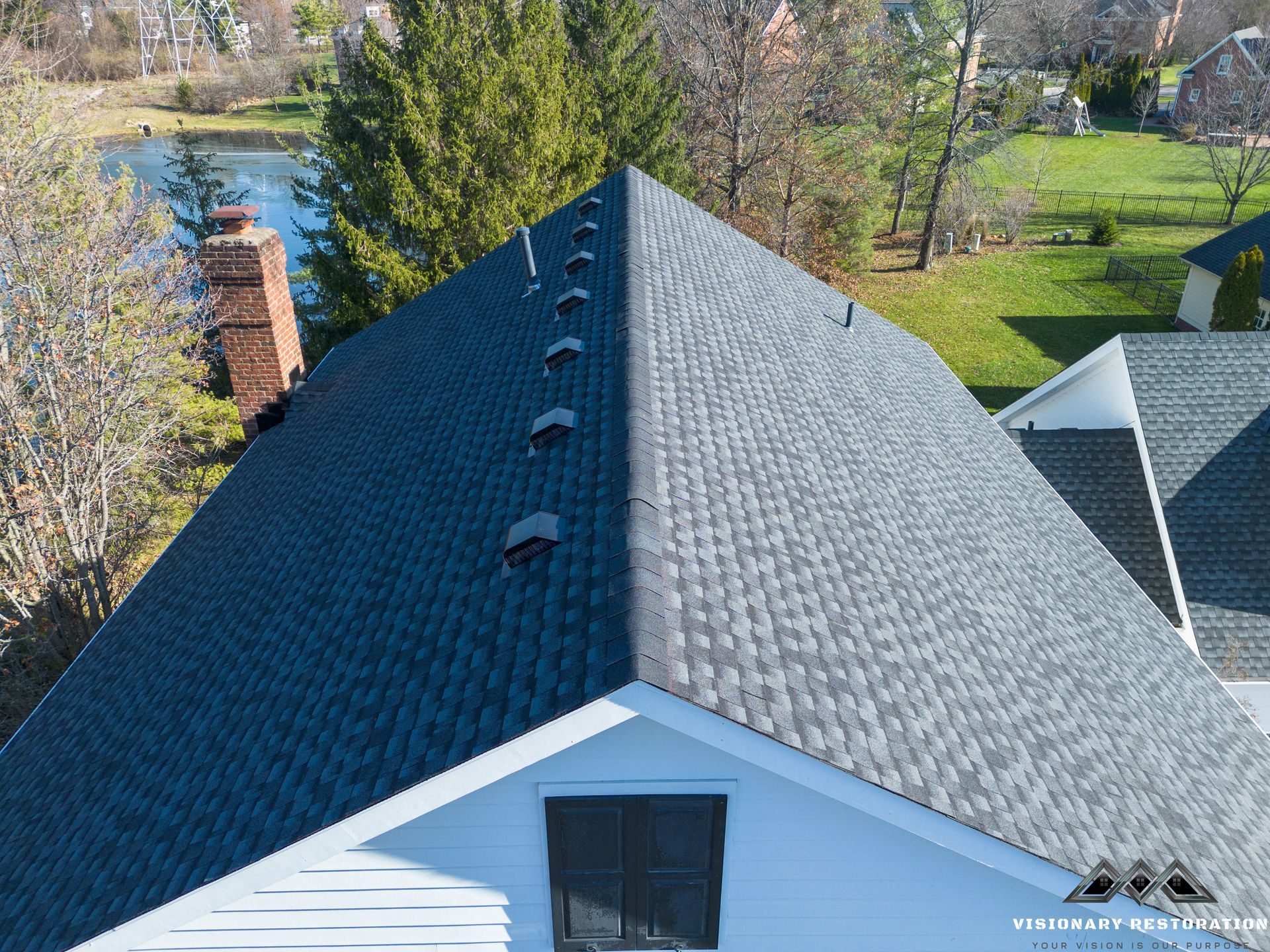 Overhead view of a dark gray shingled roof with a brick chimney and vents. White house, green yard, and lake in background.