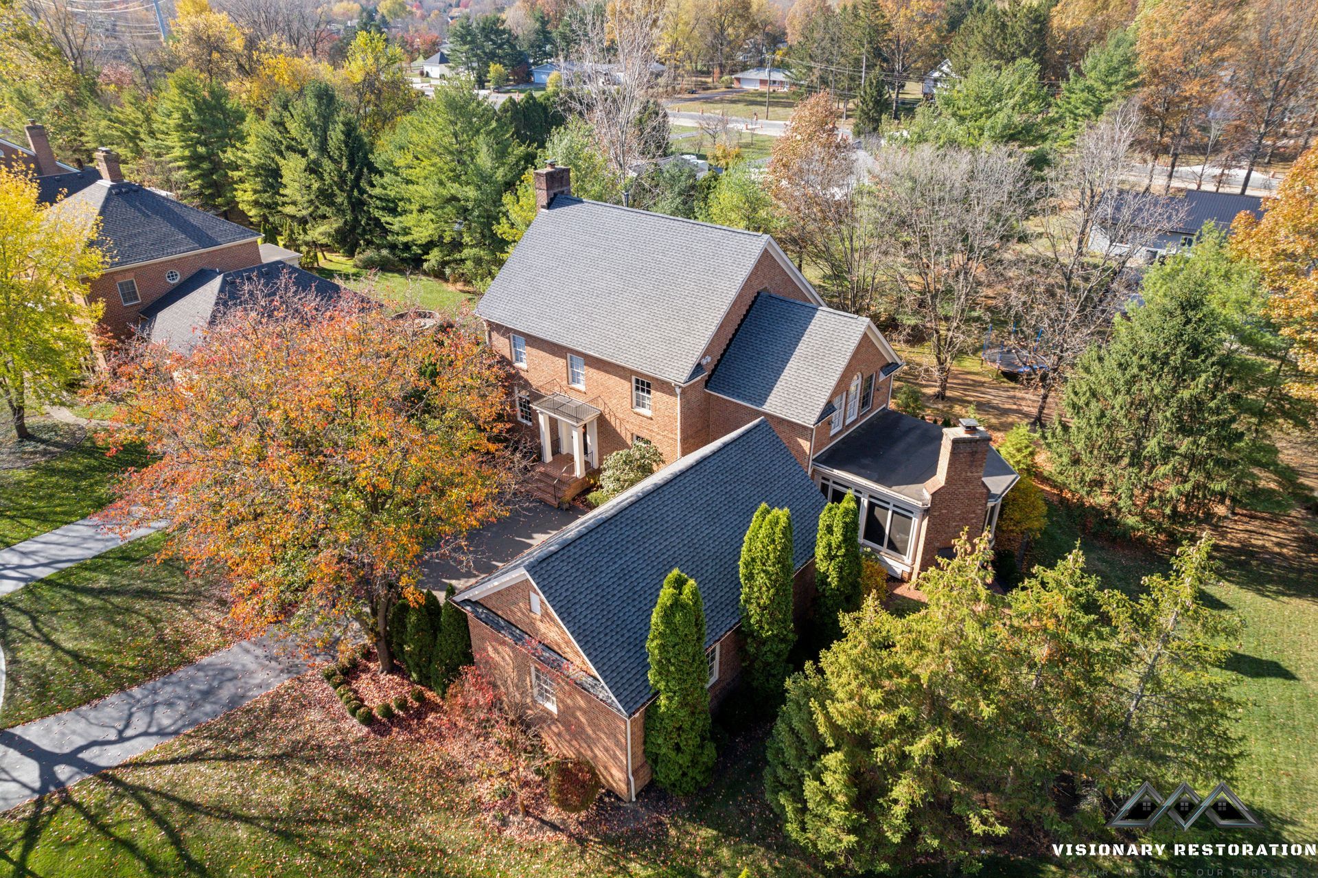 Aerial view of a brick house with a gray roof, surrounded by trees with colorful fall foliage.