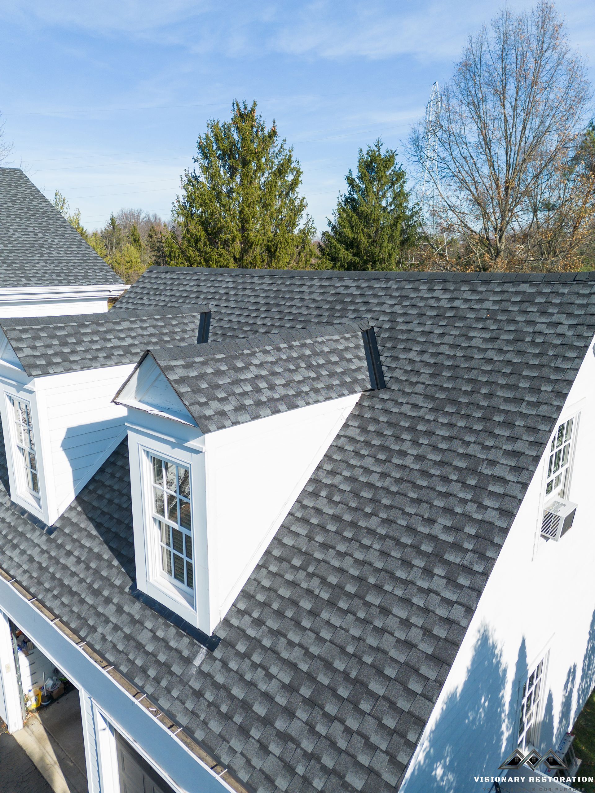 Gray asphalt shingle roof on a white house with dormer windows, under a blue sky.