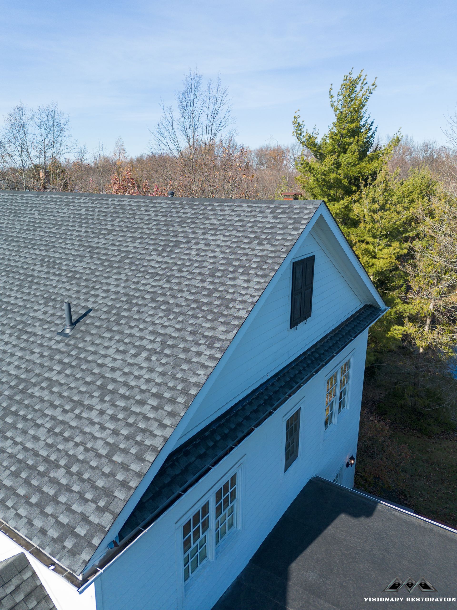 View of a house with a gray shingle roof and white siding, surrounded by trees under a blue sky.