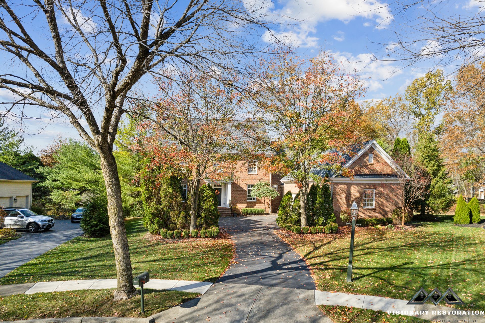 Brick house with long driveway, trees with autumn leaves, blue sky.