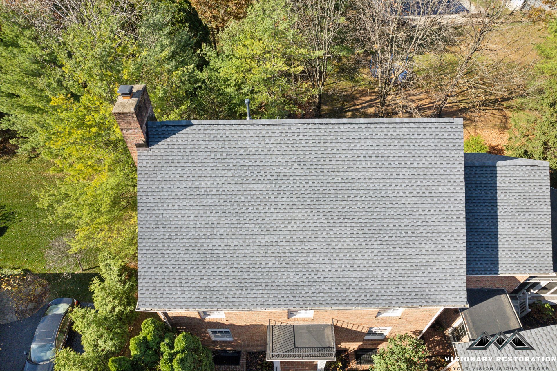 Overhead view of a gray shingle roof with a brick chimney, surrounded by green trees.