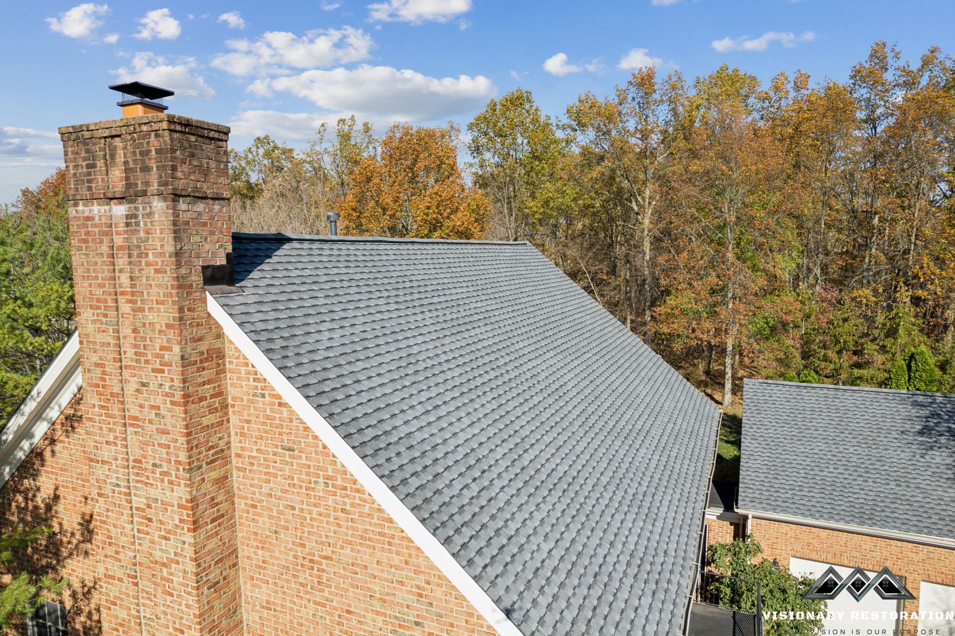 Brick house with gray shingled roof and brick chimney against a backdrop of autumn trees.