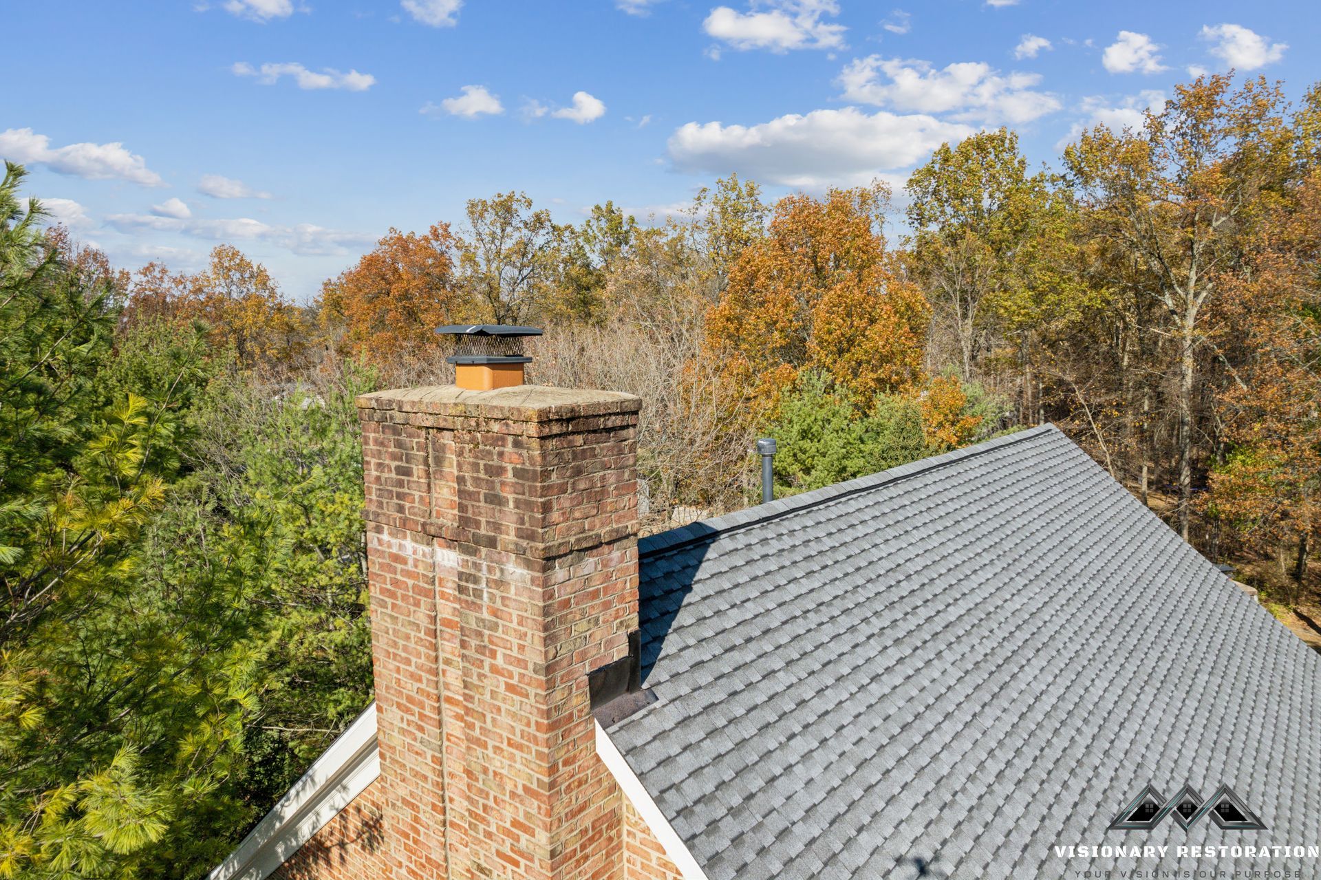 Brick chimney on a gray shingle roof, surrounded by fall foliage under a blue sky.