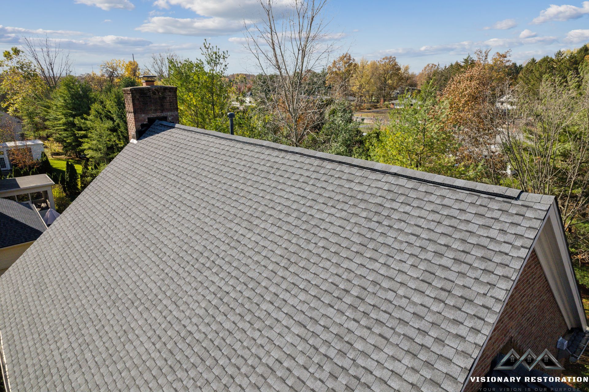 Gray shingle roof of a house with a brick chimney, trees in the background under a blue sky.