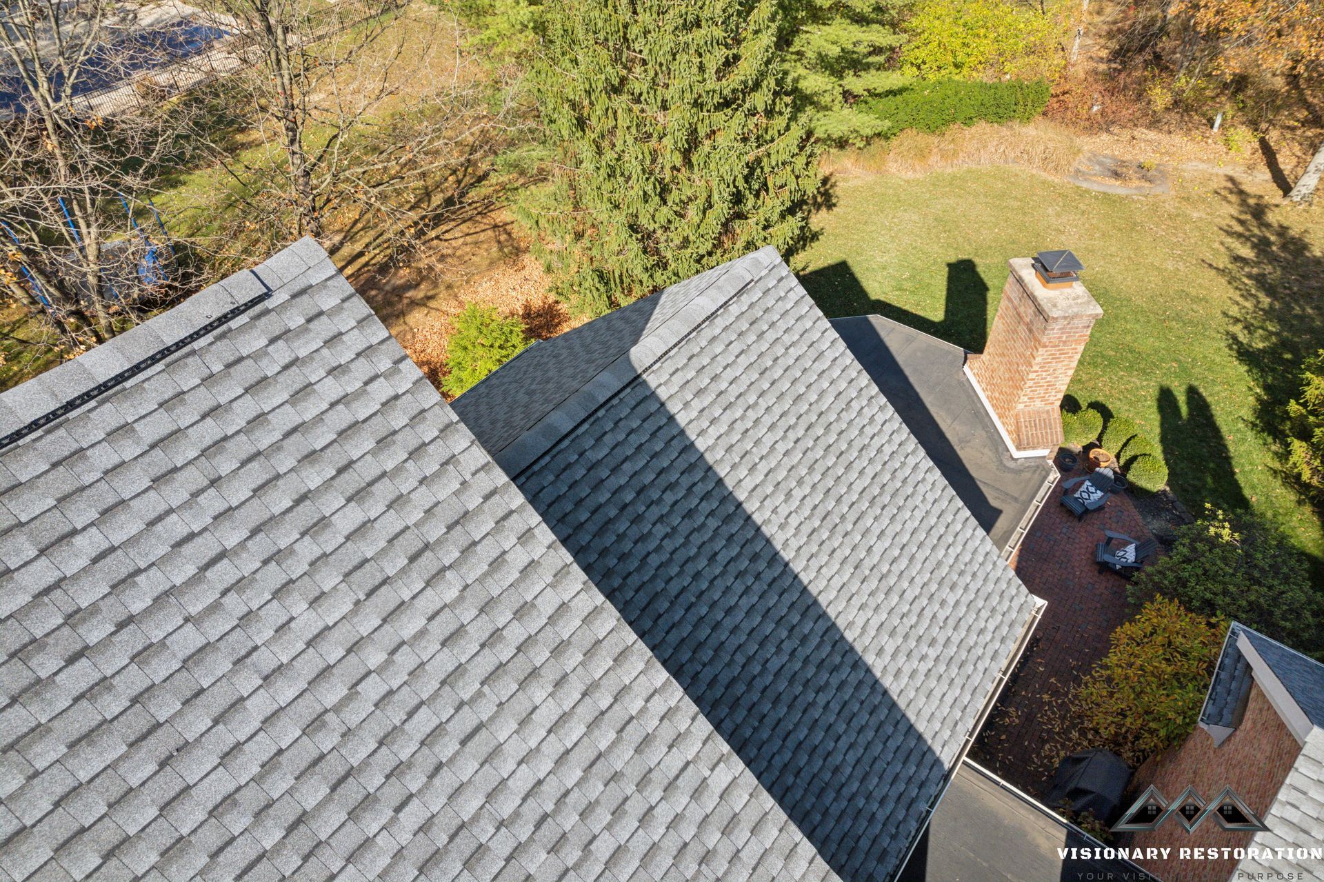 Overhead view of a house roof with gray shingles. A brick chimney and green yard are visible.