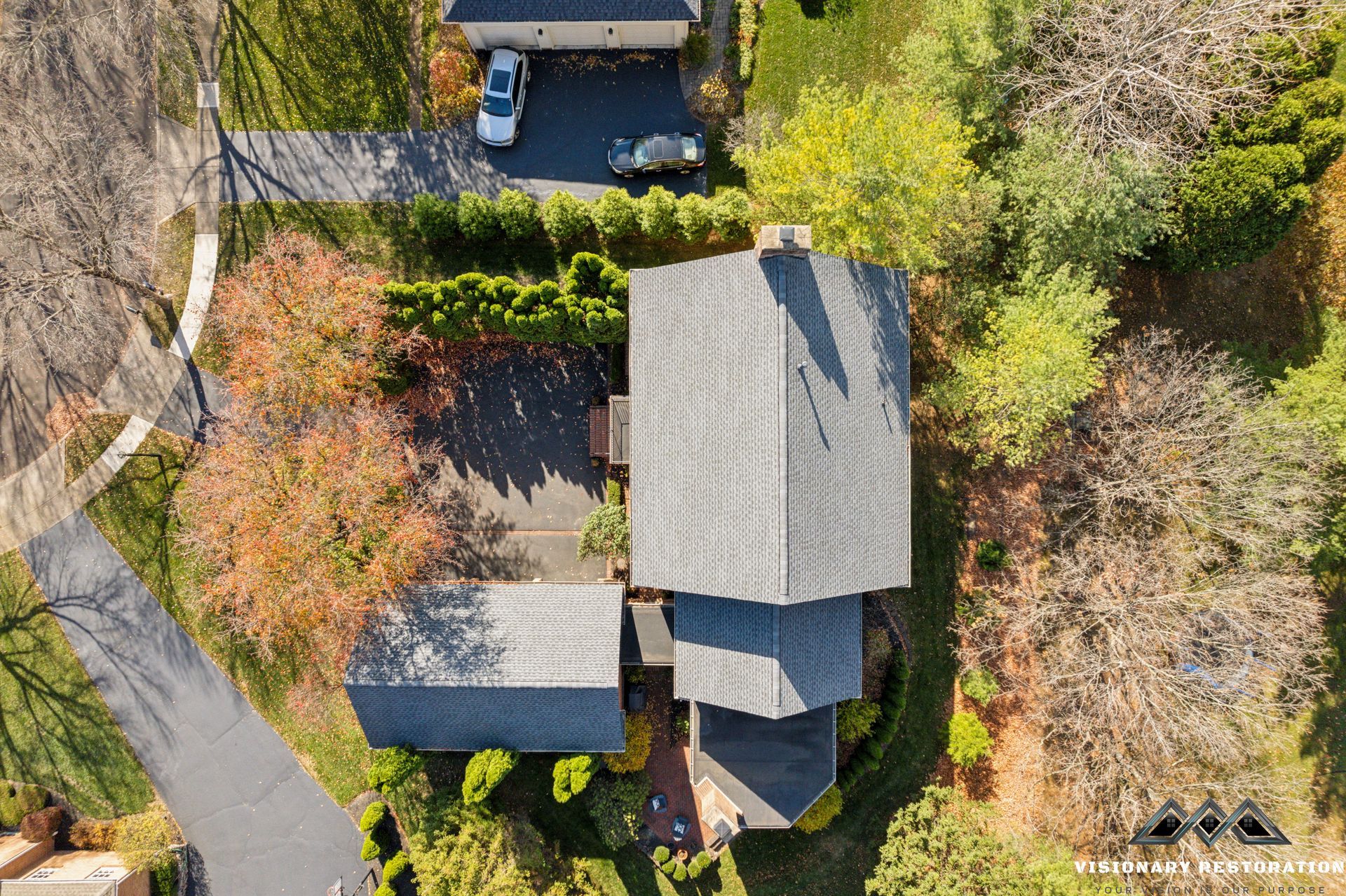 Aerial view of a house with a gray roof and two cars parked in the driveway surrounded by trees.
