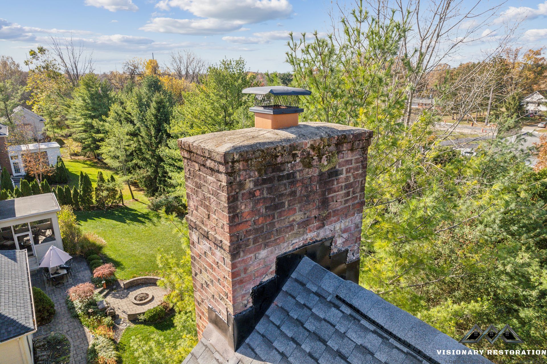 Brick chimney on a shingled roof with a metal cap, surrounded by trees and a backyard.