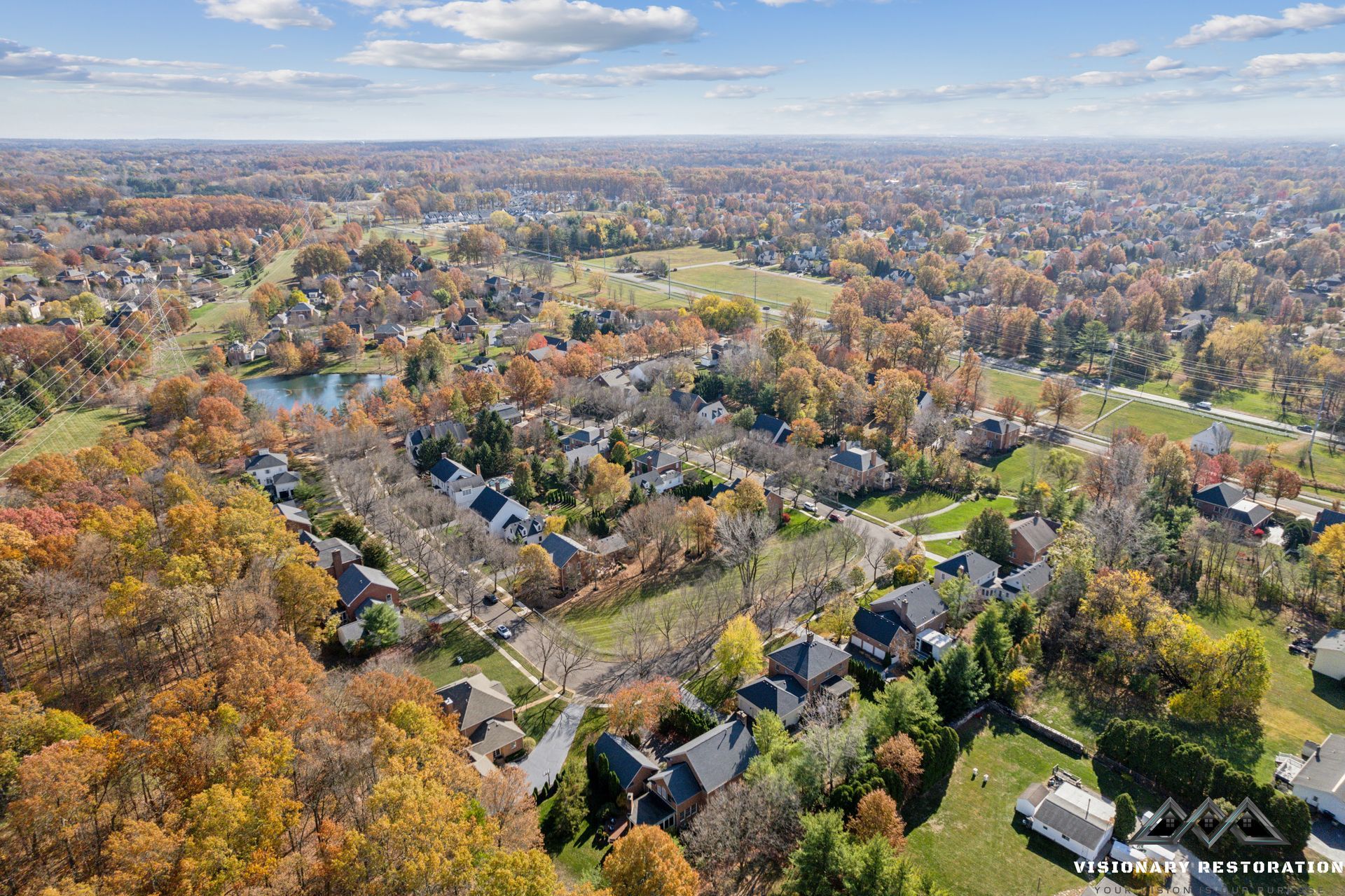 Aerial view of residential neighborhood in autumn; trees with colorful foliage surround houses and a small lake.