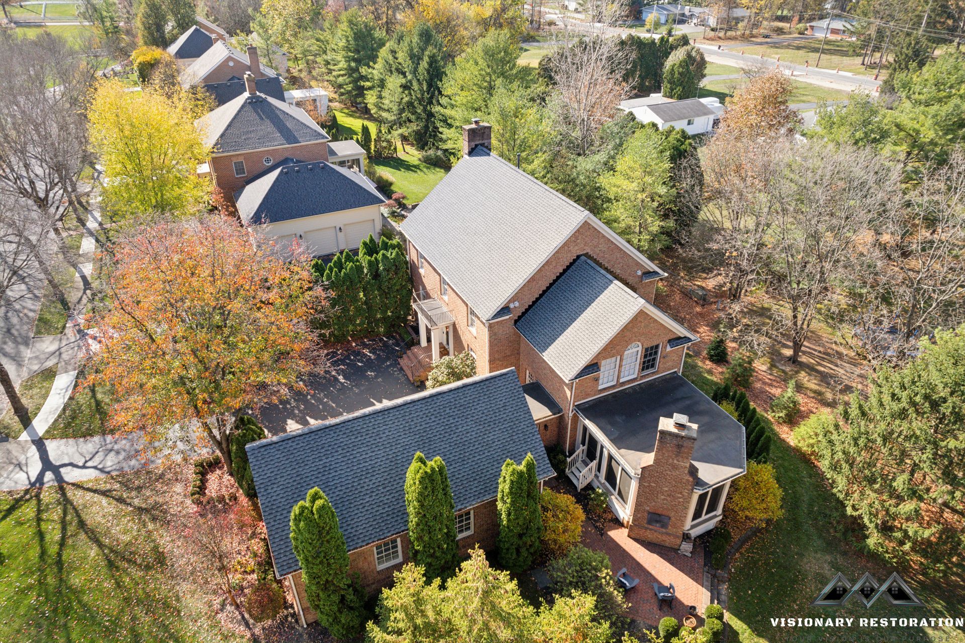 Aerial view of a brick house with a dark gray roof surrounded by trees with colorful fall foliage.