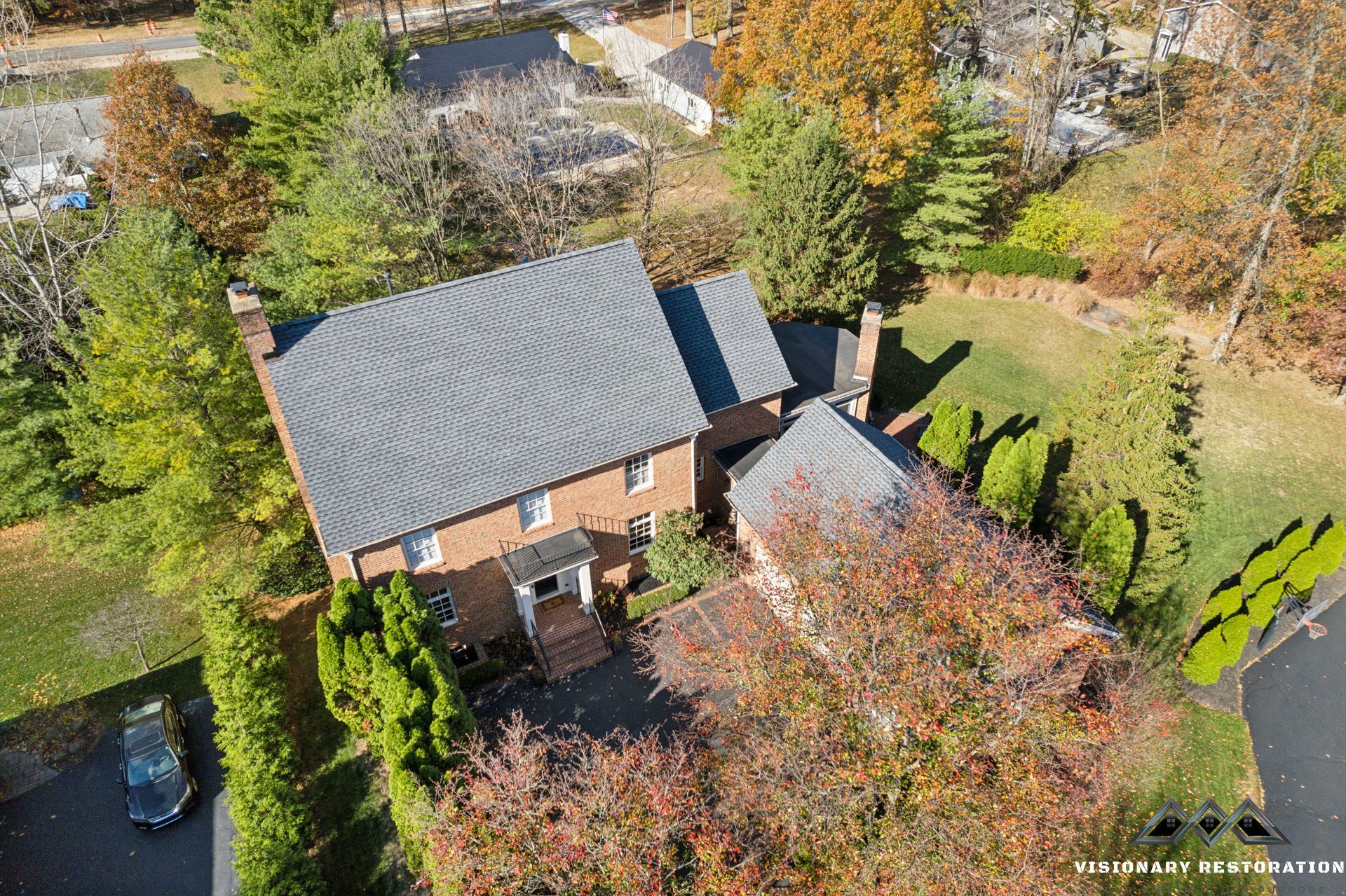 Aerial view of a brick house with a dark gray roof surrounded by trees and a driveway.